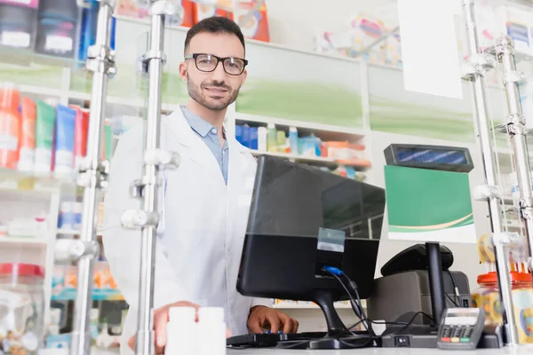 bearded pharmacist in white coat and eyeglasses smiling near computer monitor in drugstore