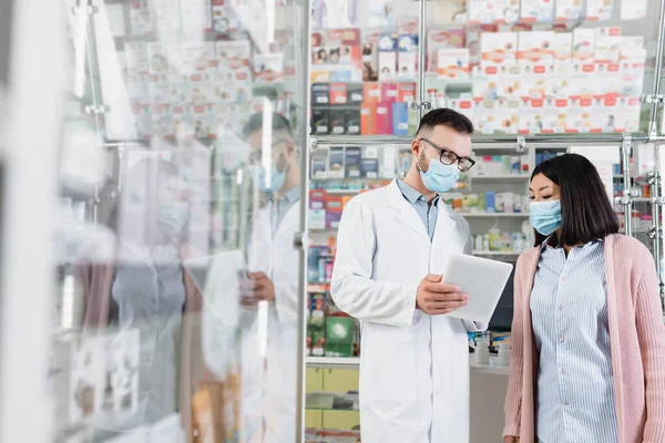 薬局でアジア人女性の近くにデジタルタブレットを持っている医療用マスクの薬剤師 — ストック写真