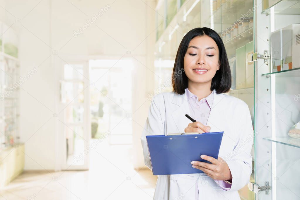 smiling asian pharmacist in white coat writing on clipboard in drugstore 