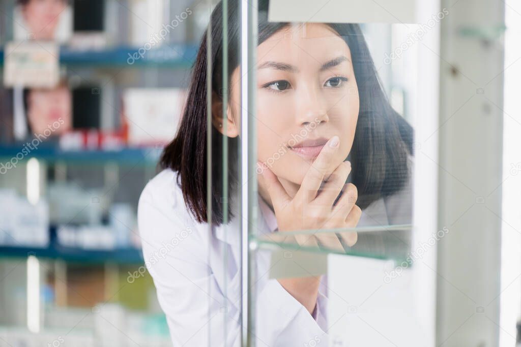pensive asian pharmacist in white coat looking at glass shelves on blurred foreground