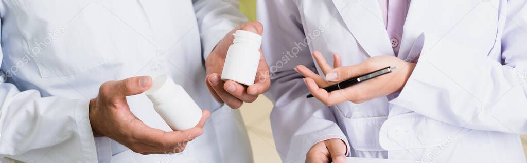 partial view of pharmacist in white coat holding bottles with medication near colleague in drugstore, banner