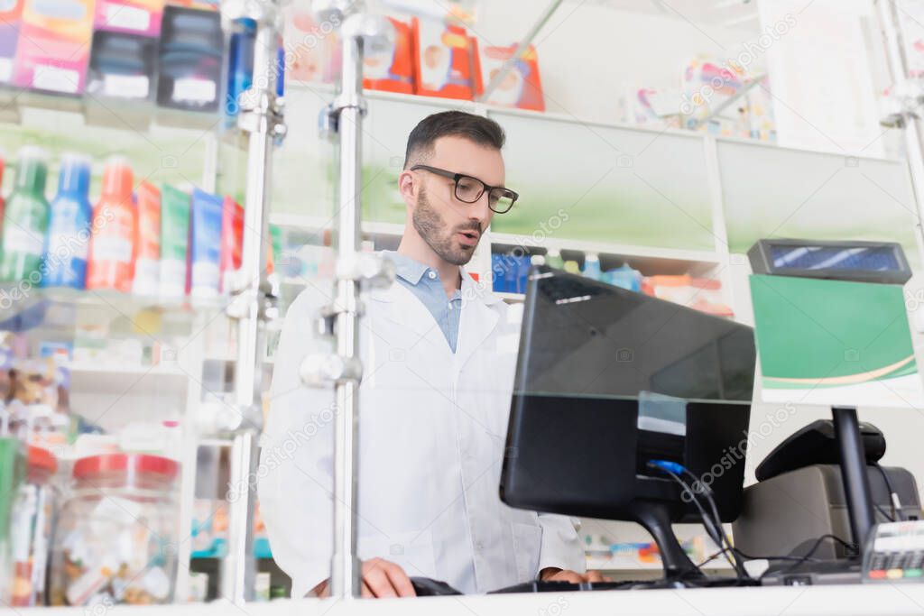 surprised pharmacist in white coat and eyeglasses looking at computer monitor in drugstore