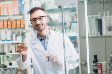 Gözlüklü neşeli eczacı elinde haplarla eczanenin cam kapısının yanında.