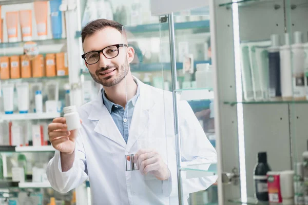 Gözlüklü neşeli eczacı elinde haplarla eczanenin cam kapısının yanında.