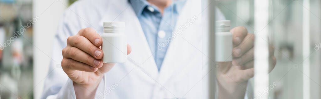 cropped view of pharmacist holding bottle with pills near glass on blurred foreground, banner