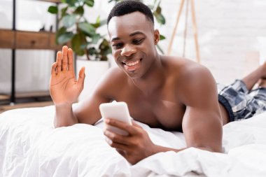 Gömleksiz Afro-Amerikalı adam video görüşmesinde el sallıyor. Akıllı telefondan yatak odasındaki bulanık ön planda. 