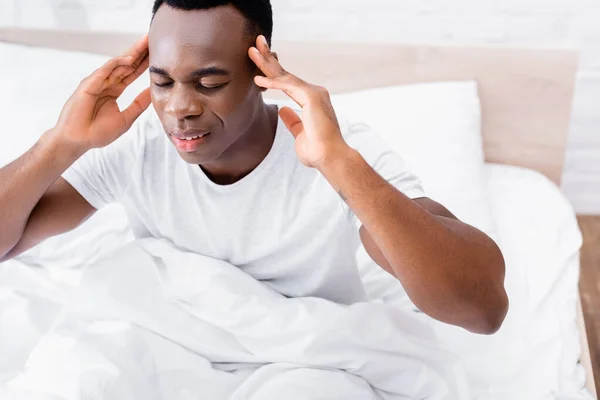 African american man with closed eyes suffering from headache on bed on blurred background