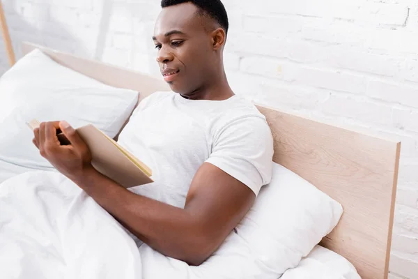 Sorridente Uomo Africano Americano Leggere Libro Sul Letto Con Biancheria — Foto Stock