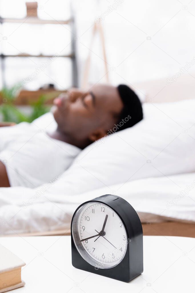 Clock and book on bedside table near african american man sleeping on blurred background 