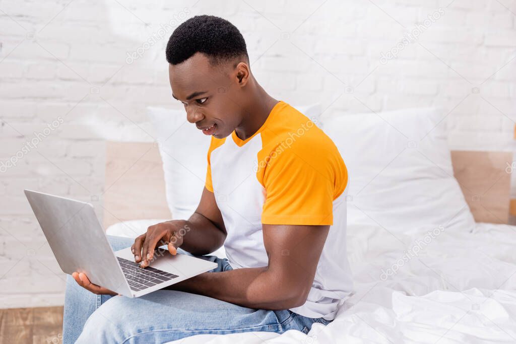 Positive african american man using laptop while sitting on bed at home 