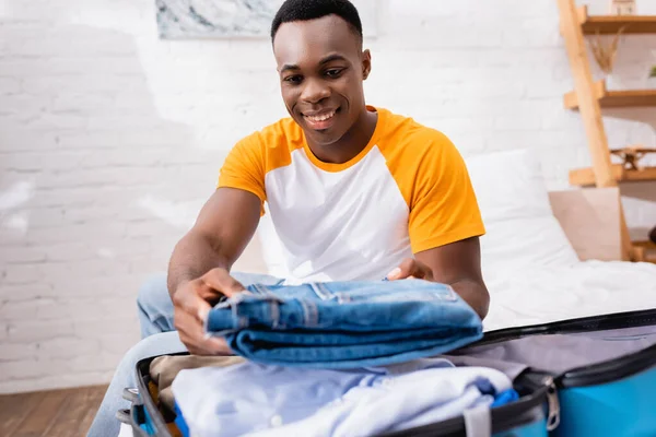 Glimlachende Afro Amerikaanse Man Zet Jeans Koffer Wazig Voorgrond Thuis — Stockfoto