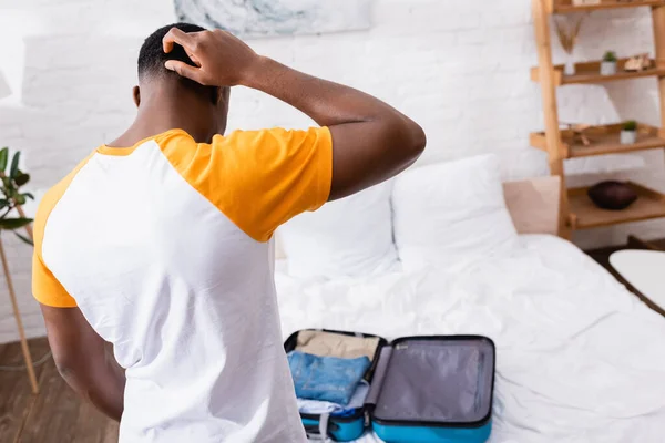 Back view of pensive african american man standing near suitcase on bed on blurred background
