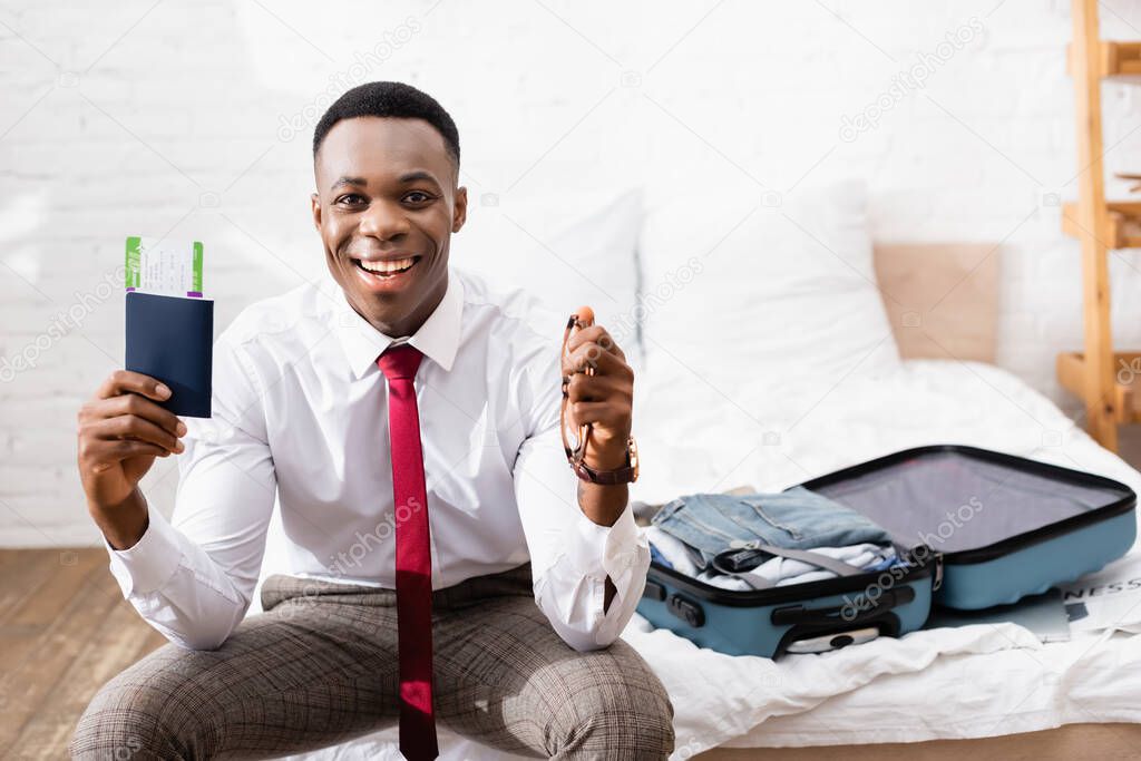 Cheerful african american businessman holding passport, air ticket and eyeglasses near suitcase on blurred background on bed 