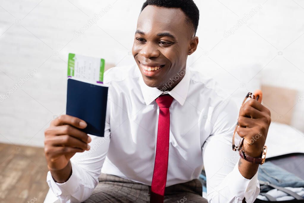 Smiling african american businessman holding eyeglasses and passport with air ticket on blurred foreground 