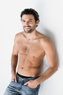 Kot pantolonlu, beyaz arka planda kameraya bakan neşeli gömleksiz adam.
