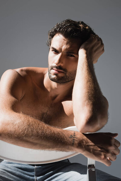 Shirtless man in jeans looking at camera while sitting on chair isolated on grey 