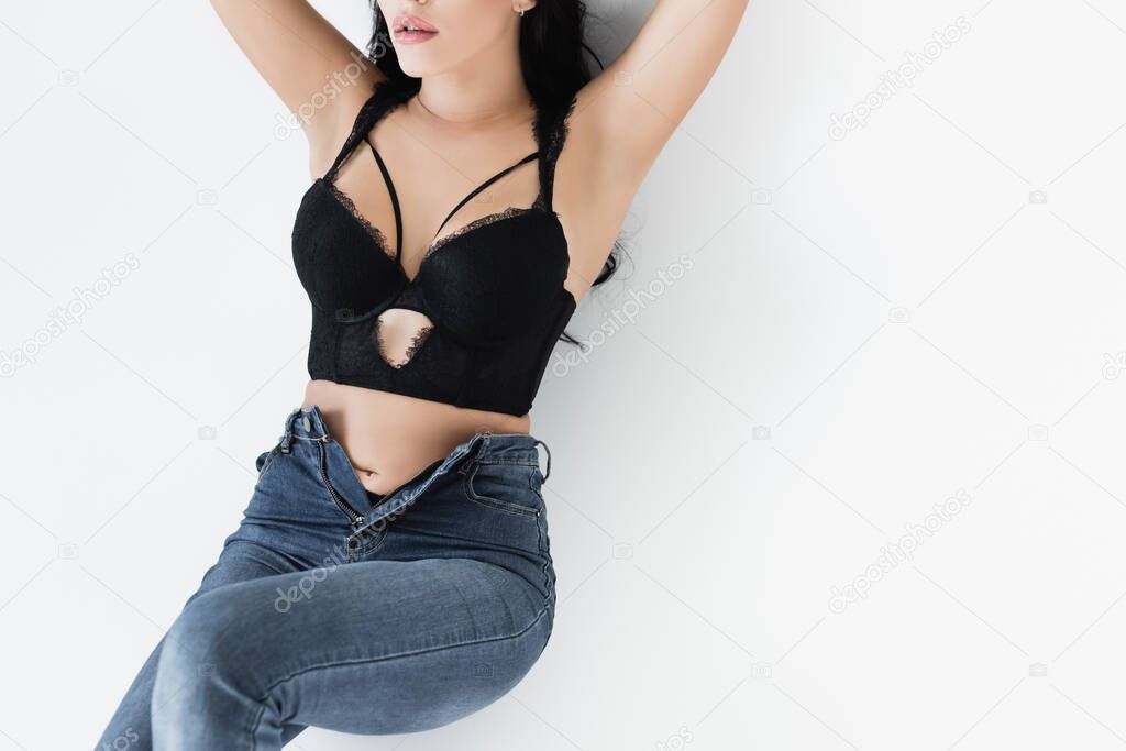 Cropped view of sensual woman in bra and jeans lying on white background