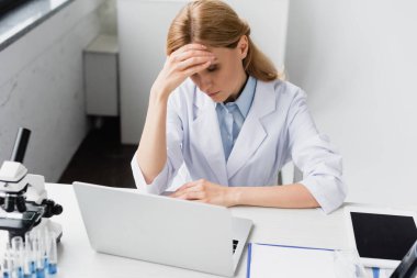 Laptop ve mikroskop yanında yüzünü örten beyaz önlüklü üzgün bir bilim adamı.