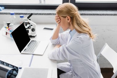 Beyaz önlüklü üzgün bilim adamı laptopun yanında boş ekran ve masasında mikroskop ile yüzünü kapatıyor.