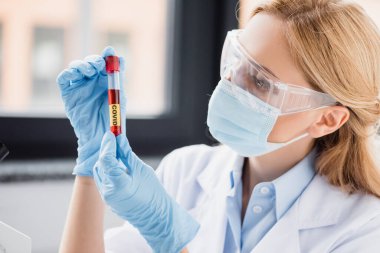 Tıbbi maskeli ve gözlüklü bilim adamı laboratuarında renkli harflerle test tüpüne bakıyor.