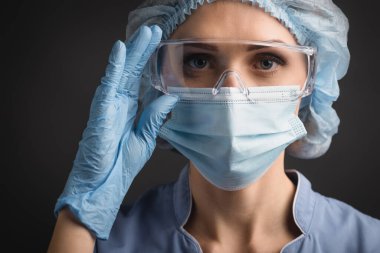 Mavi şapkalı ve maskeli hemşire koyu gri kameraya bakarken gözlükleri ayarlıyor.