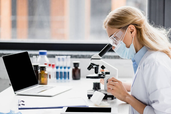 scientist in medical mask and goggles looking through microscope near gadgets with blank screen 