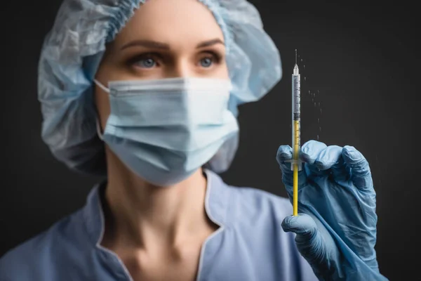 暗灰色で隔離された背景のぼやけた背景の医療用マスクの看護師の手にワクチンスプラッシュ注射器 — ストック写真