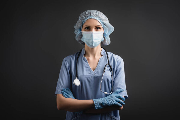 nurse in medical mask and latex gloves standing with crossed arms isolated on dark grey 