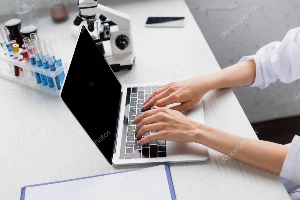 cropped view of scientist typing on laptop with blank screen near microscope on desk