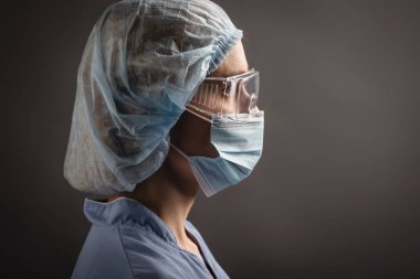 Tıbbi şapkalı, maskeli ve gözlüklü hemşirenin yan görünüşü koyu griye bakıyor.