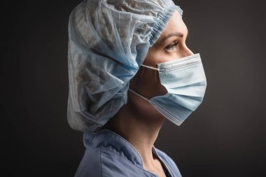Tıbbi şapkalı ve maskeli hemşirenin yan görünüşü koyu griye bakıyor. 
