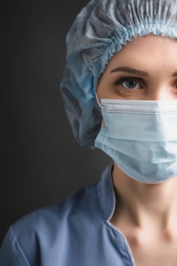 Tıbbi şapkalı ve maskeli hemşirenin koyu gri renkli kameraya bakarken kısmi görüntüsü