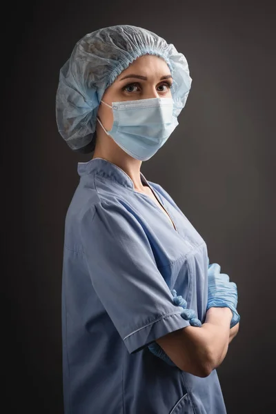 nurse in medical cap and mask standing with crossed arms and looking at camera isolated on dark grey