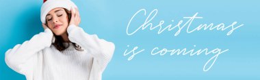 young brunette woman with closed eyes adjusting hat near christmas is coming lettering on blue, banner clipart