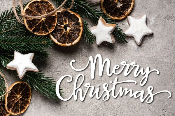 Top view of pine branch with dried orange pieces and cookies near merry christmas lettering on grey background
