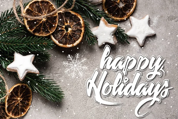 Top view of pine branch with dried orange pieces and cookies near happy holidays lettering on grey background