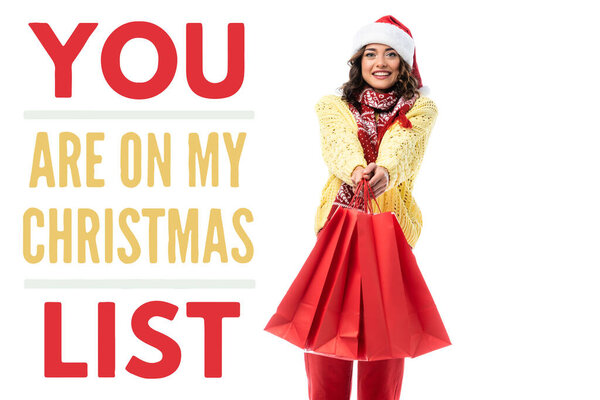joyful young woman in santa hat and scarf with ornament holding red shopping bags near you are on my christmas list lettering on white 