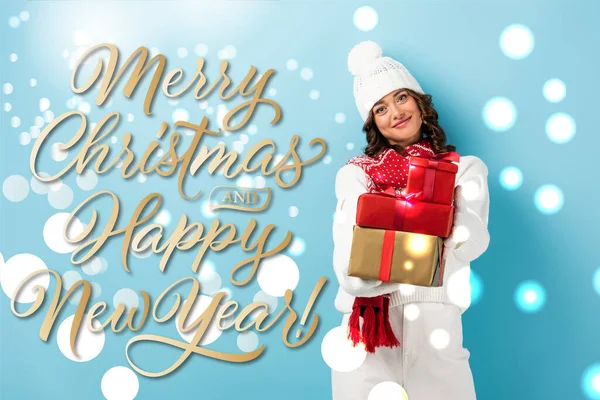 young joyful woman in winter outfit holding wrapped presents near merry christmas and happy new year lettering on blue