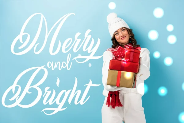 young joyful woman in winter outfit holding wrapped presents near merry and bright lettering on blue