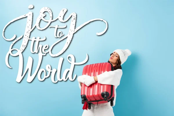 young woman in winter outfit carrying heavy suitcase near joy to the world lettering on blue