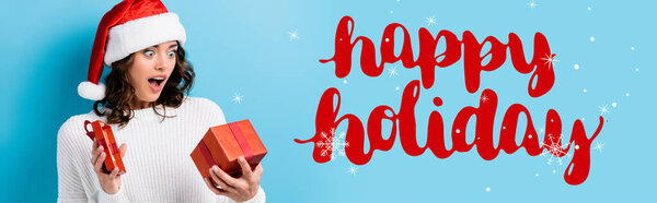 young shocked woman looking at gift box near happy holidays lettering on blue, banner