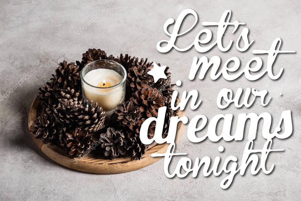 Scented candle with pine cones on wooden plate near lets meet in our dreams tonight  on grey background