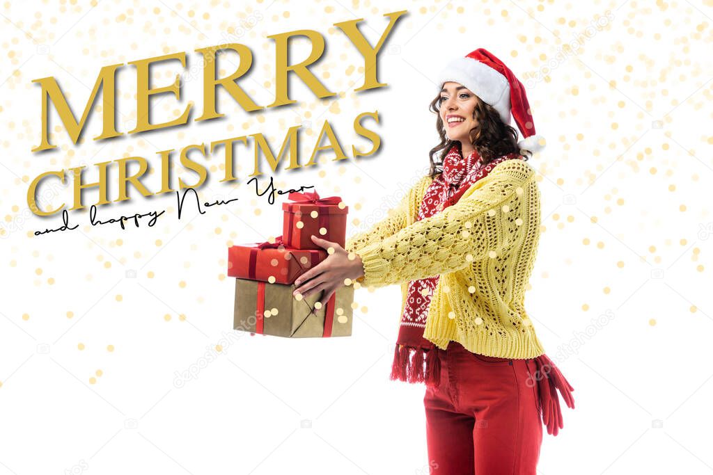 joyful young woman in santa hat, scarf and knitted sweater holding gifts near merry christmas and happy new year lettering on white