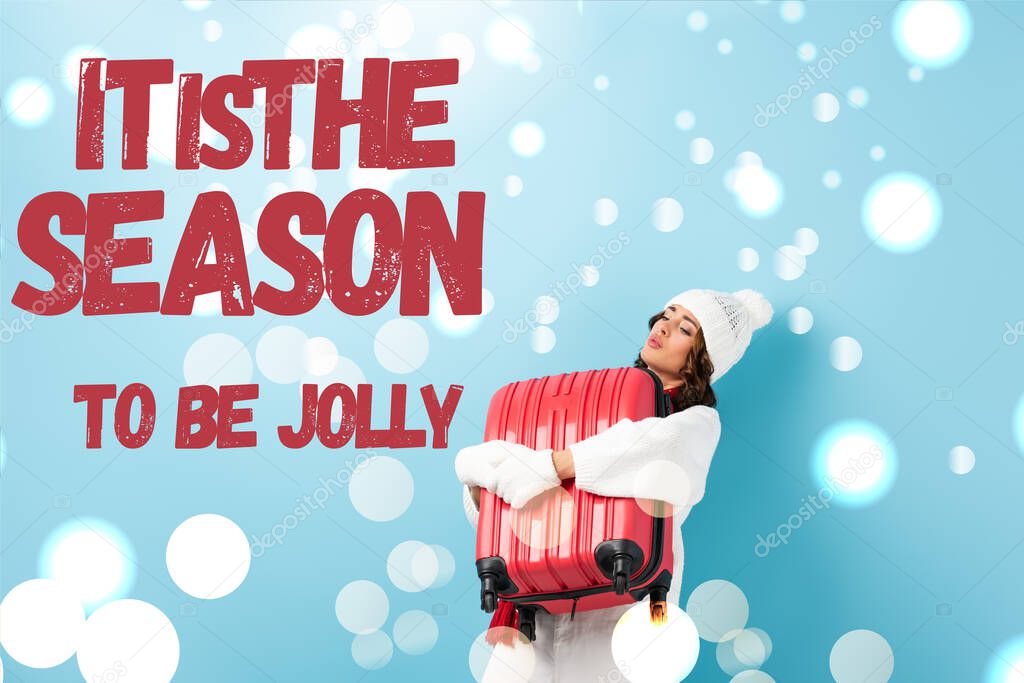 young woman in winter outfit carrying heavy suitcase near it is the season to be jolly lettering on blue