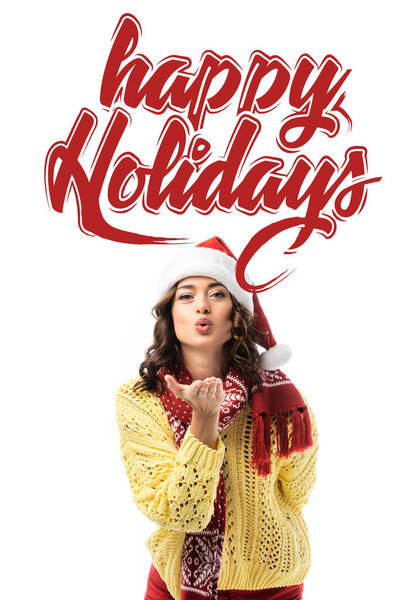 young woman in santa hat and scarf sending air kiss near happy holidays lettering on white 