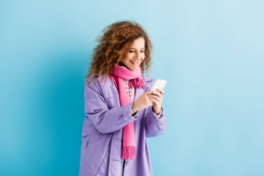Kışlık ceketli ve pembe örgü atkılı mutlu genç bir kadın mavi üzerine akıllı telefondan mesaj atıyor.