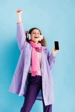 Kablosuz kulaklıklar, kışlık palto ve pembe örgü fular takan mavi ekranda boş ekranlı akıllı telefon tutan heyecanlı genç kadın.