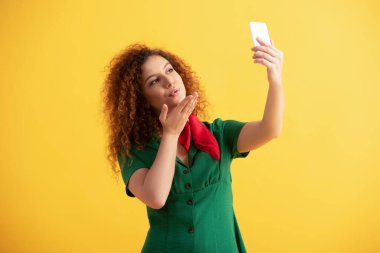 Yeşil elbiseli kıvırcık kadın hava öpücüğü gönderirken sarıda selfie çekiyor. 