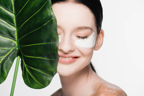 smiling young beautiful woman with vitiligo and eye patches on face near green leaf isolated on white