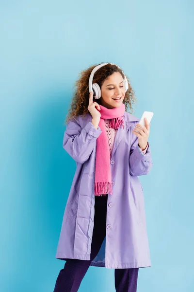 happy young woman in wireless headphones, winter coat and pink knitted scarf using smartphone on blue